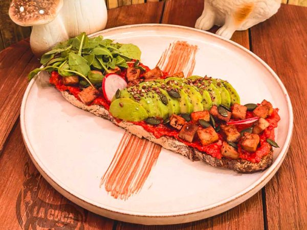 LA VEGANA: Tostada vegana de hummus de pimiento rojo asado y nueces, tofu a la plancha marinado, aguacate, rúcula, semillas y pipas de calabaza.