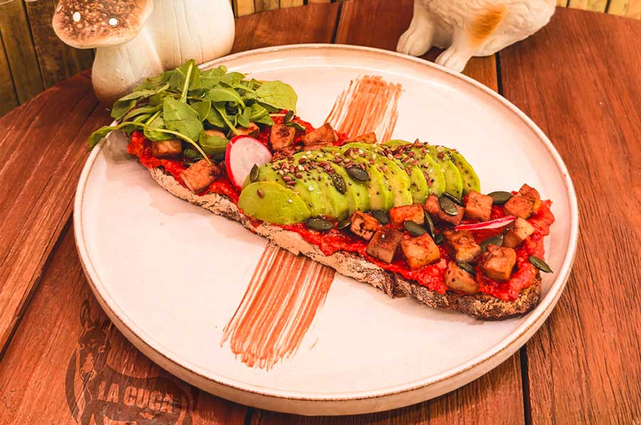 LA VEGANA: Tostada vegana de hummus de pimiento rojo asado y nueces, tofu a la plancha marinado, aguacate, rúcula, semillas y pipas de calabaza.